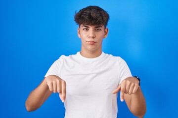 Hispanic teenager standing over blue background pointing down looking sad and upset, indicating...