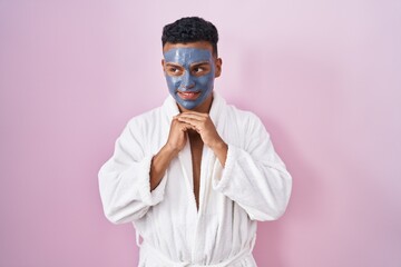 Young hispanic man wearing beauty face mask and bath robe laughing nervous and excited with hands...