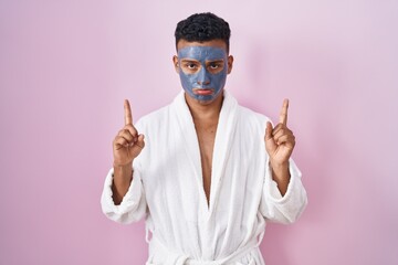 Young hispanic man wearing beauty face mask and bath robe pointing up looking sad and upset, indicating direction with fingers, unhappy and depressed.