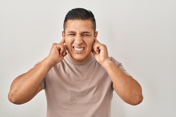 Fototapeta premium Hispanic young man standing over white background covering ears with fingers with annoyed expression for the noise of loud music. deaf concept.