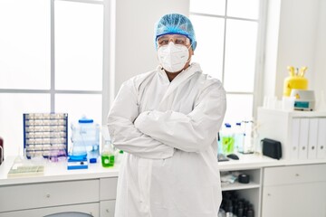 Young latin man scientist wearing covid protection uniform standing with arms crossed gesture at laboratory