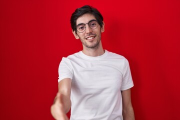 Obraz premium Young hispanic man standing over red background smiling friendly offering handshake as greeting and welcoming. successful business.