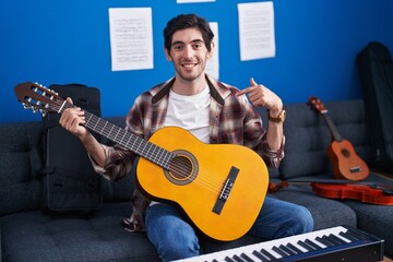 Young hispanic man playing classic guitar at music studio pointing finger to one self smiling happy and proud