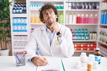 Hispanic young man working at pharmacy drugstore with hand on chin thinking about question, pensive expression. smiling with thoughtful face. doubt concept.
