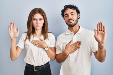 Young couple wearing casual clothes standing together swearing with hand on chest and open palm,...