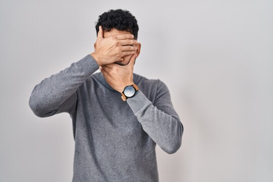 Hispanic man with beard standing over white background covering eyes and mouth with hands, surprised and shocked. hiding emotion