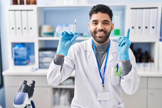 Hispanic man with beard working at scientist laboratory holding syringe smiling happy pointing with hand and finger to the side - Powered by Adobe