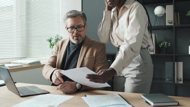 Mid-shot of senior Caucasian man typing on laptop while his African American female colleague bringing him two CVs. He then asking question about them, his colleague answering, leaving