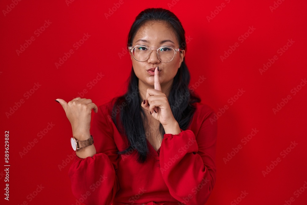 Canvas Prints Asian young woman standing over red background asking to be quiet with finger on lips pointing with hand to the side. silence and secret concept.