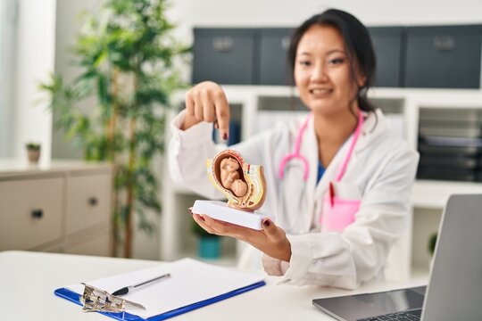 Young chinese woman wearing doctor uniform holding anatomical model of uterus with fetus at clinic