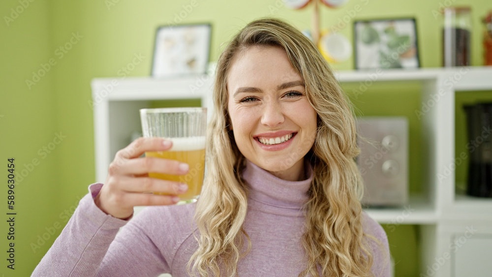 Sticker Young blonde woman holding glass of orange juice sitting on table at dinning room