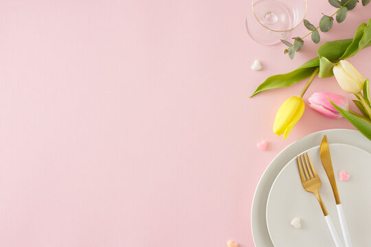 Women's Day celebration concept. Flat lay photo of white plate cutlery knife fork empty glass bunch of colorful tulips and small hearts baubles on pastel pink background with copy space