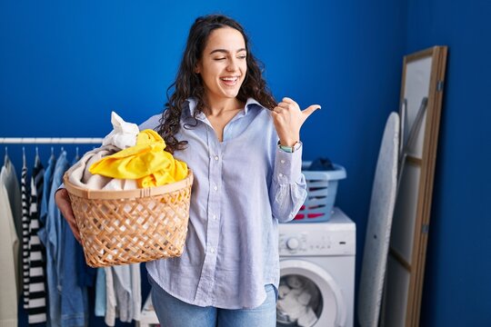 Young brunette woman holding laundry basket pointing thumb up to the side smiling happy with open mouth