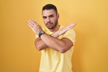 Young hispanic man standing over yellow background rejection expression crossing arms doing...
