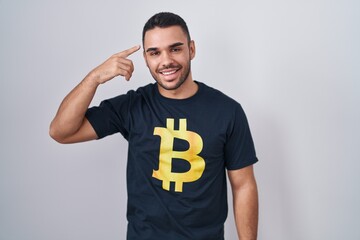 Young hispanic man wearing bitcoin t shirt smiling pointing to head with one finger, great idea or thought, good memory