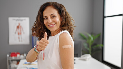 Middle age hispanic woman doing ok gesture standing with band aid on arm at clinic