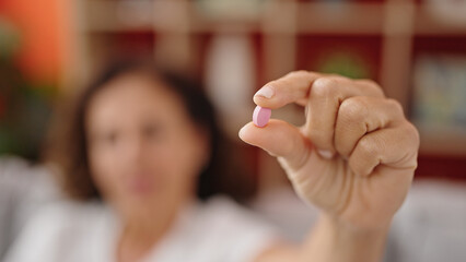 Middle age hispanic woman sitting on sofa holding pill at home