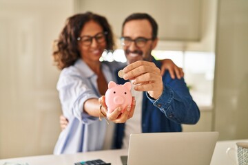 Middle age hispanic couple smiling confident saving coin on piggy bank at kitchen