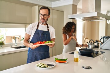 Middle age couple cooking mediterranean food at home in shock face, looking skeptical and sarcastic, surprised with open mouth