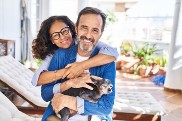 Middle age hispanic couple hugging each other sitting on hammock with dog at terrace
