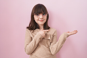 Little hispanic girl wearing glasses amazed and smiling to the camera while presenting with hand and pointing with finger.