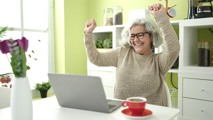 Middle age woman with grey hair using laptop sitting on table dancing at home