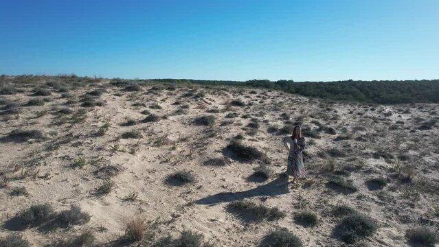 A girl walks in the dune singing with her ceremonial drum with the view of the forest behind her