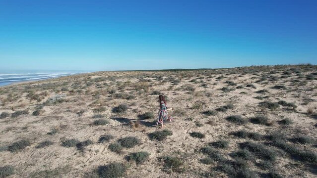 A woman in colorful ceremony dress running in the dune between the beach and the forest with her drum