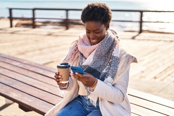 Obraz premium African american woman using smartphone drinking coffee sitting on bench at seaside