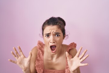 Young teenager girl standing over pink background crazy and mad shouting and yelling with aggressive expression and arms raised. frustration concept.