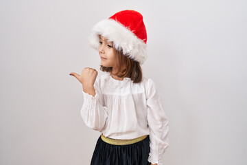 Little hispanic girl wearing christmas hat pointing thumb up to the side smiling happy with open mouth