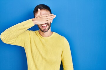 Hispanic man standing over blue background smiling and laughing with hand on face covering eyes for surprise. blind concept.