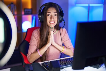 Young hispanic woman playing video games praying with hands together asking for forgiveness smiling confident.