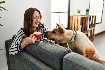 Young hispanic woman drinking coffee sitting on sofa with dogs at home