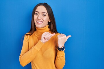 Young brunette woman standing over blue background pointing to the back behind with hand and thumbs up, smiling confident