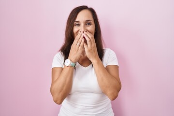 Middle age brunette woman standing over pink background laughing and embarrassed giggle covering...