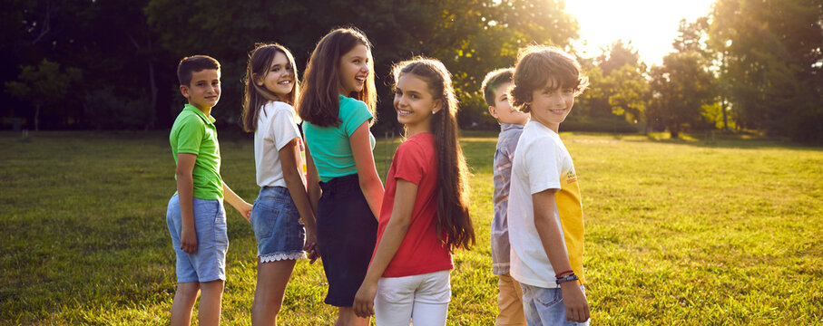 Children friendship. Group of cheerful and cute preteen friends walking together in park on sunny summer day. Smiling boys and girls in casual clothes having fun outdoors. Panoramic web banner.