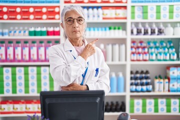 Middle age woman with tattoos working at pharmacy drugstore pointing with hand finger to the side showing advertisement, serious and calm face