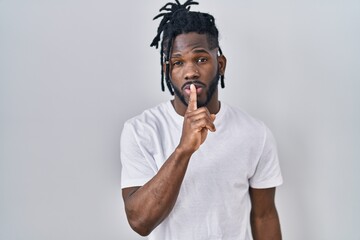 African man with dreadlocks wearing casual t shirt over white background asking to be quiet with finger on lips. silence and secret concept.