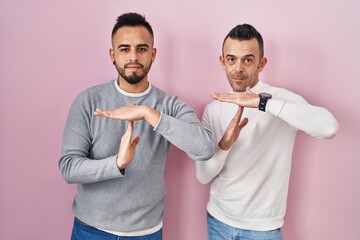 Homosexual couple standing over pink background doing time out gesture with hands, frustrated and...
