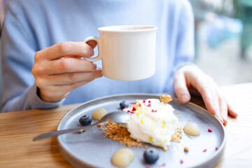 A woman's hand with a white cup close-up, a blue plate with dessert and berries. A girl in a cafe is treated to a delicious dessert. A woman in a blue sweater sits at a wooden table