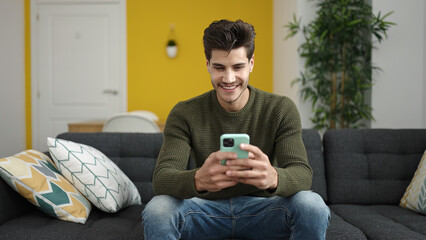 Young hispanic man using smartphone sitting on sofa at home