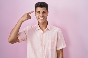 Obraz premium Young hispanic man standing over pink background smiling pointing to head with one finger, great idea or thought, good memory