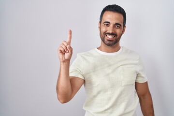 Fototapeta premium Hispanic man with beard standing over isolated background showing and pointing up with finger number one while smiling confident and happy.