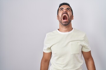 Hispanic man with beard standing over isolated background angry and mad screaming frustrated and furious, shouting with anger. rage and aggressive concept.