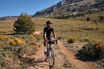 Fototapeta premium Man riding gravel bike on gravel road in mountains with scenic view.Cyclist riding on gravel bike.Cyclist practicing on gravel road.Man cyclist wearing cycling kit and helmet.Alicante region in Spain.
