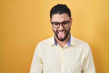 Hispanic young man wearing business clothes and glasses sticking tongue out happy with funny expression. emotion concept.