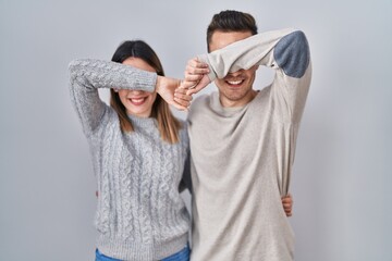 Young hispanic couple standing over white background smiling cheerful playing peek a boo with hands...