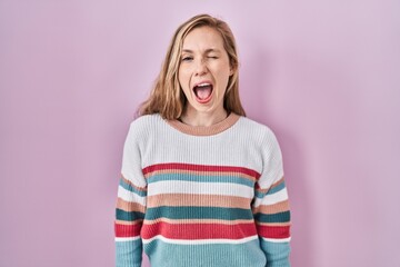 Young blonde woman standing over pink background winking looking at the camera with sexy...
