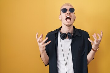 Young caucasian man wearing sunglasses standing over yellow background crazy and mad shouting and yelling with aggressive expression and arms raised. frustration concept.